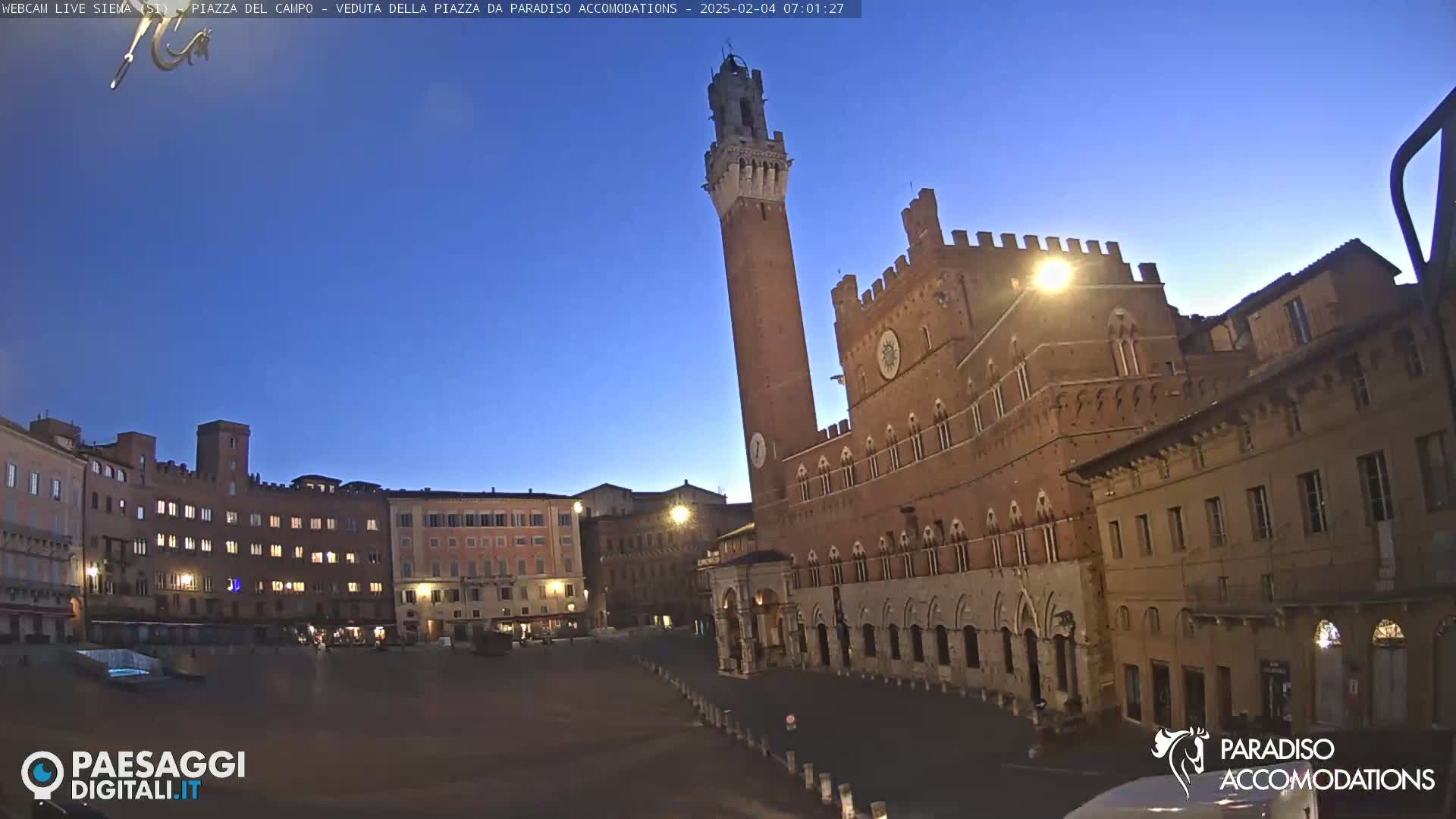 A large, brick public square is shown at twilight, dominated by a tall, slender tower and a large, ornate building.