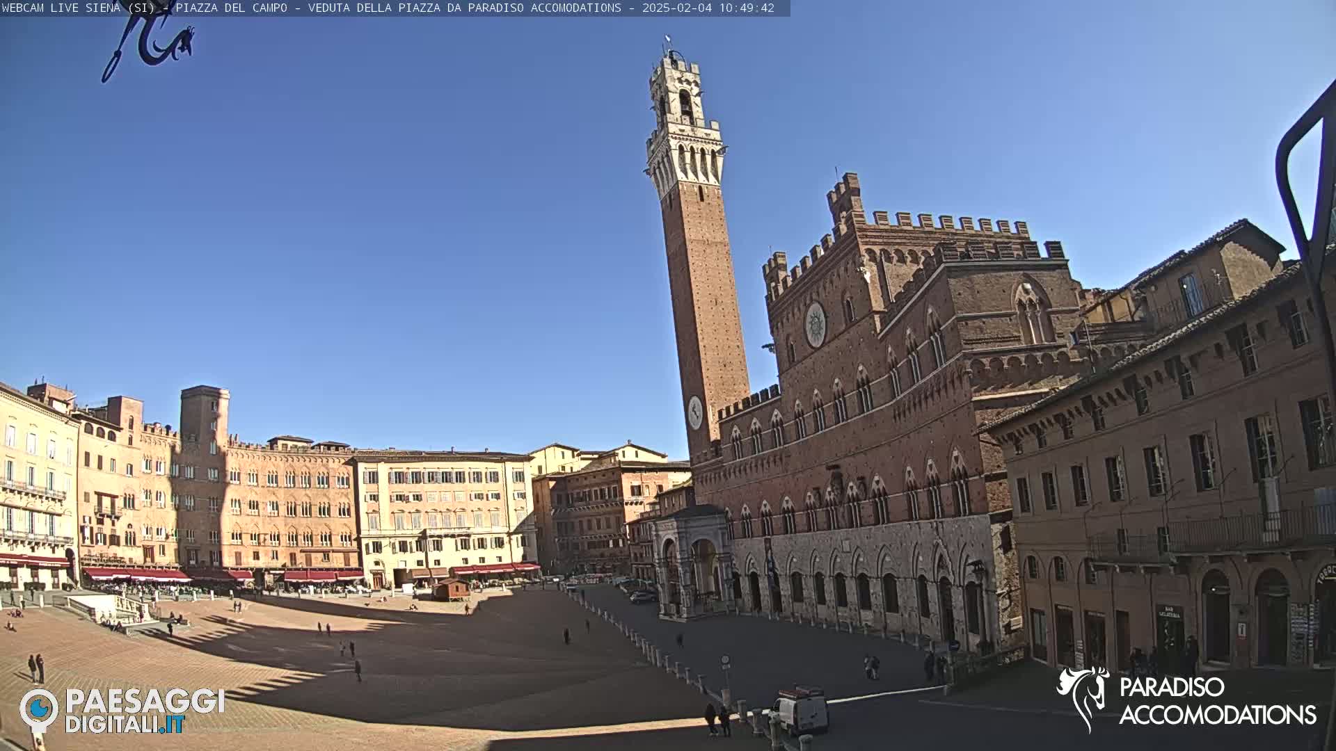 A large, brick public square in Italy is mostly empty under a clear blue sky, with a tall tower and ornate buildings surrounding it.