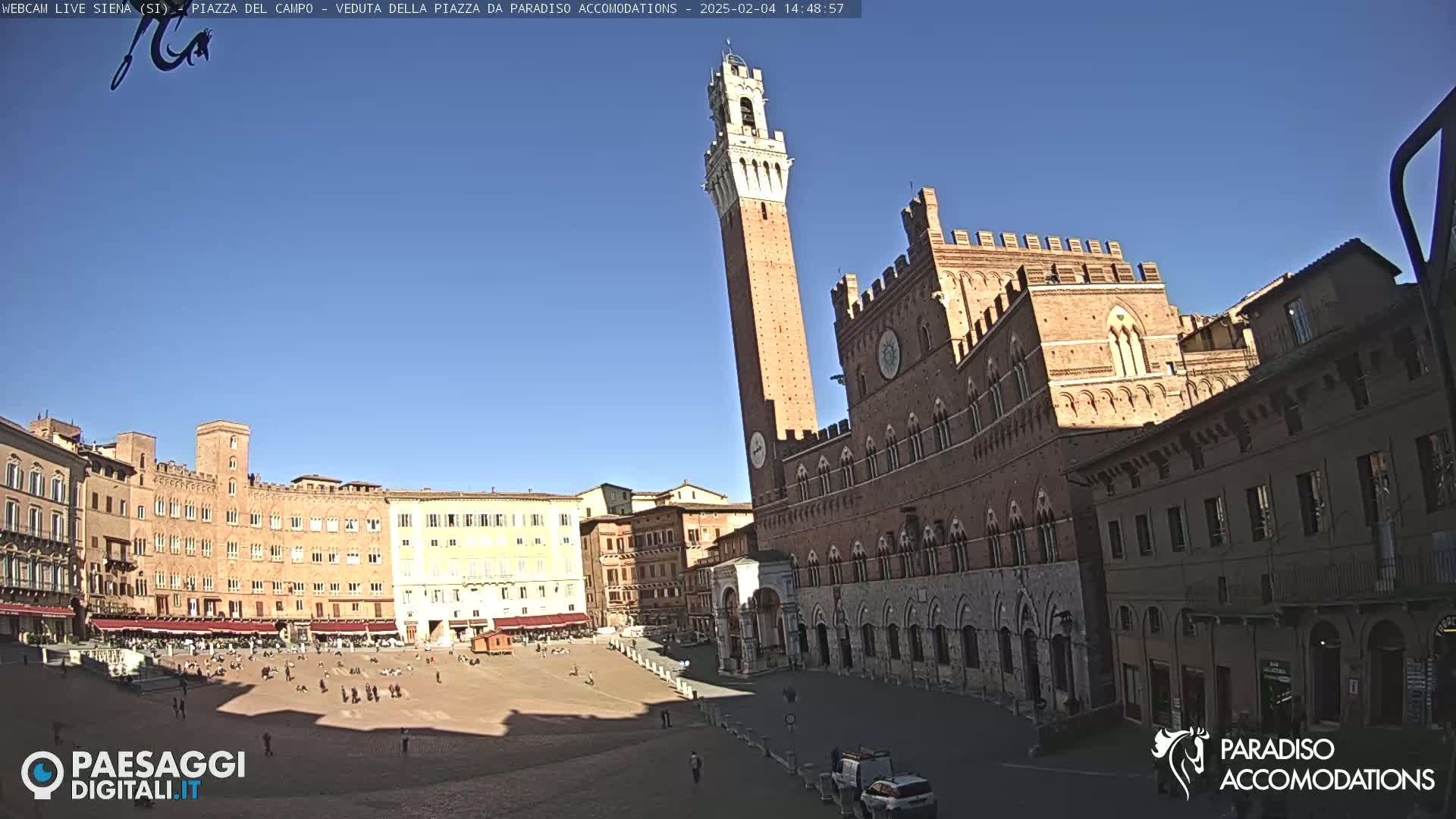 A large, brick piazza in Italy is shown on a sunny day, with a tall, slender tower and a large palace visible.
