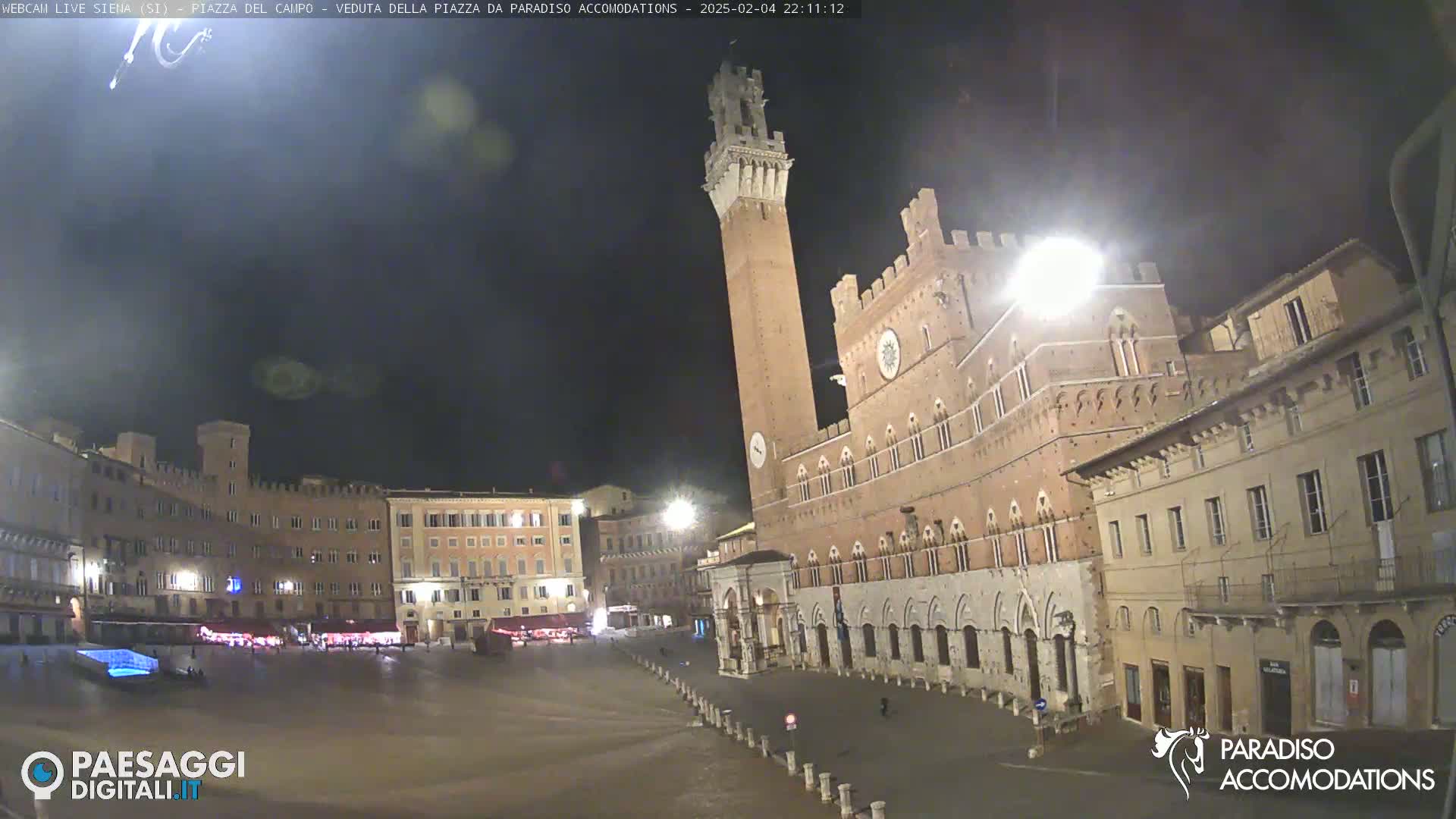 A nighttime view of a large, mostly empty town square in Italy, surrounded by historic brick buildings, including a tall tower.