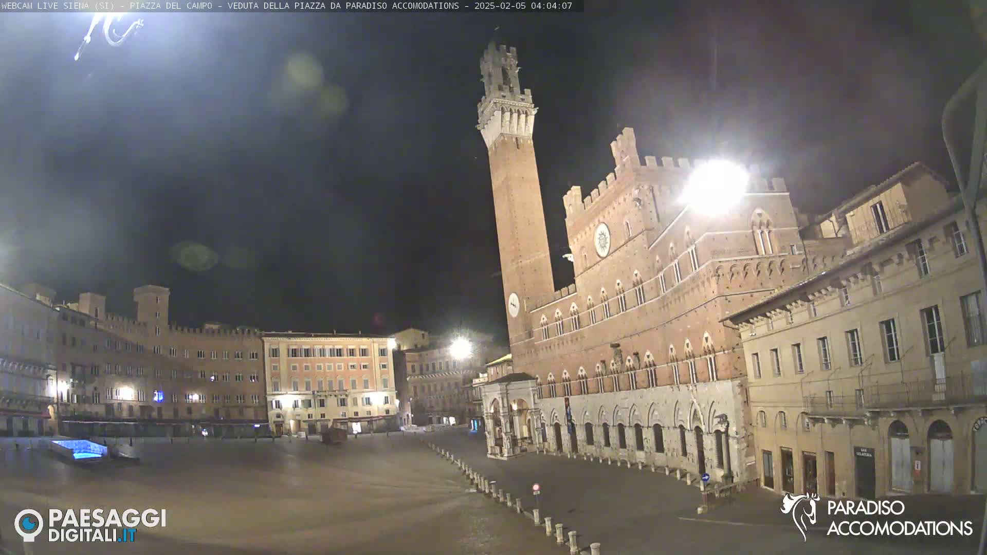 A mostly empty, cobblestone plaza at night is surrounded by brick buildings, including a tall tower and a large palace, under a dark sky.
