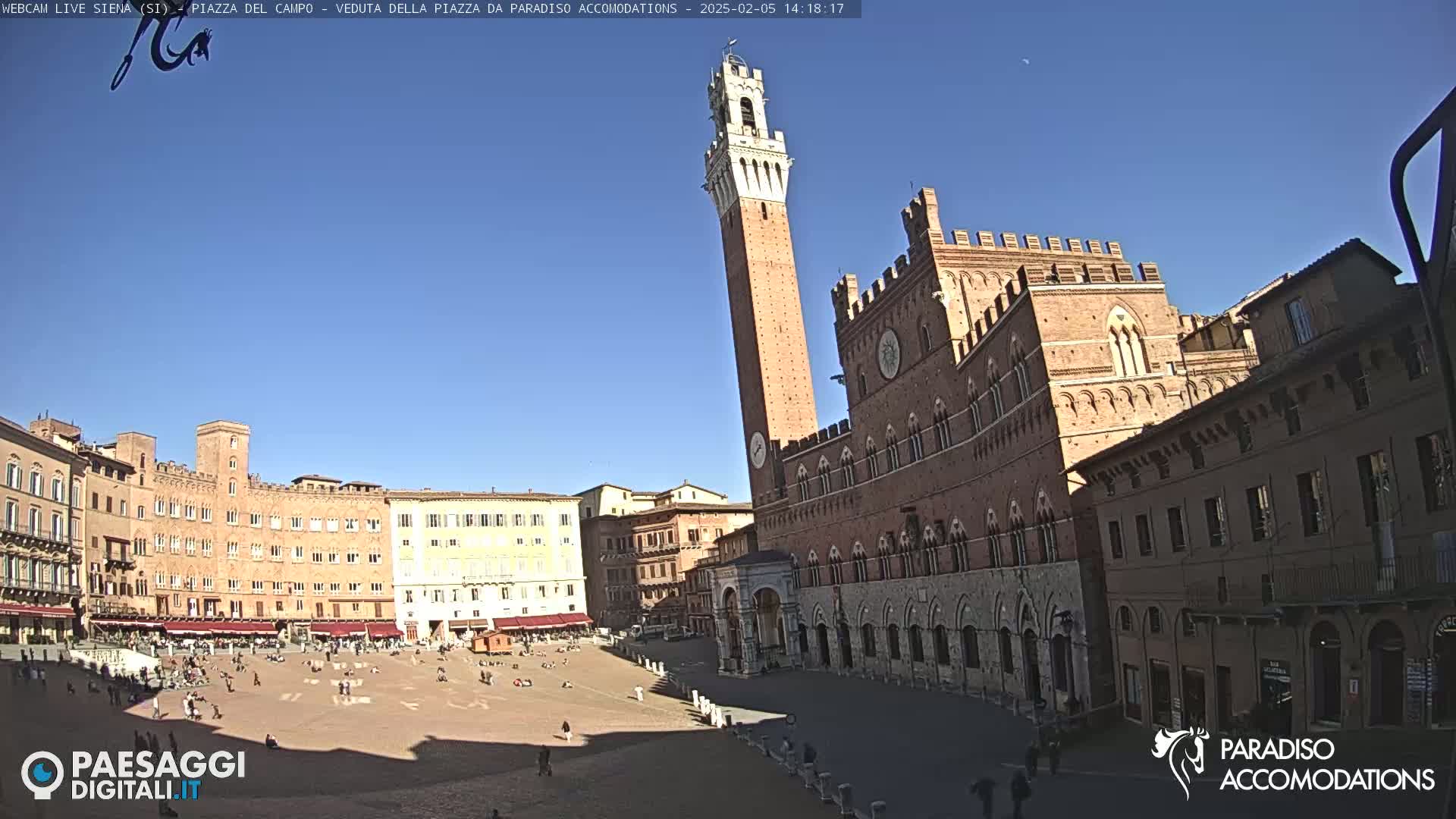 A large, brick piazza in Siena, Italy, is shown on a sunny day with a tall tower and several historic buildings surrounding it, and a few people are sparsely scattered across the square.