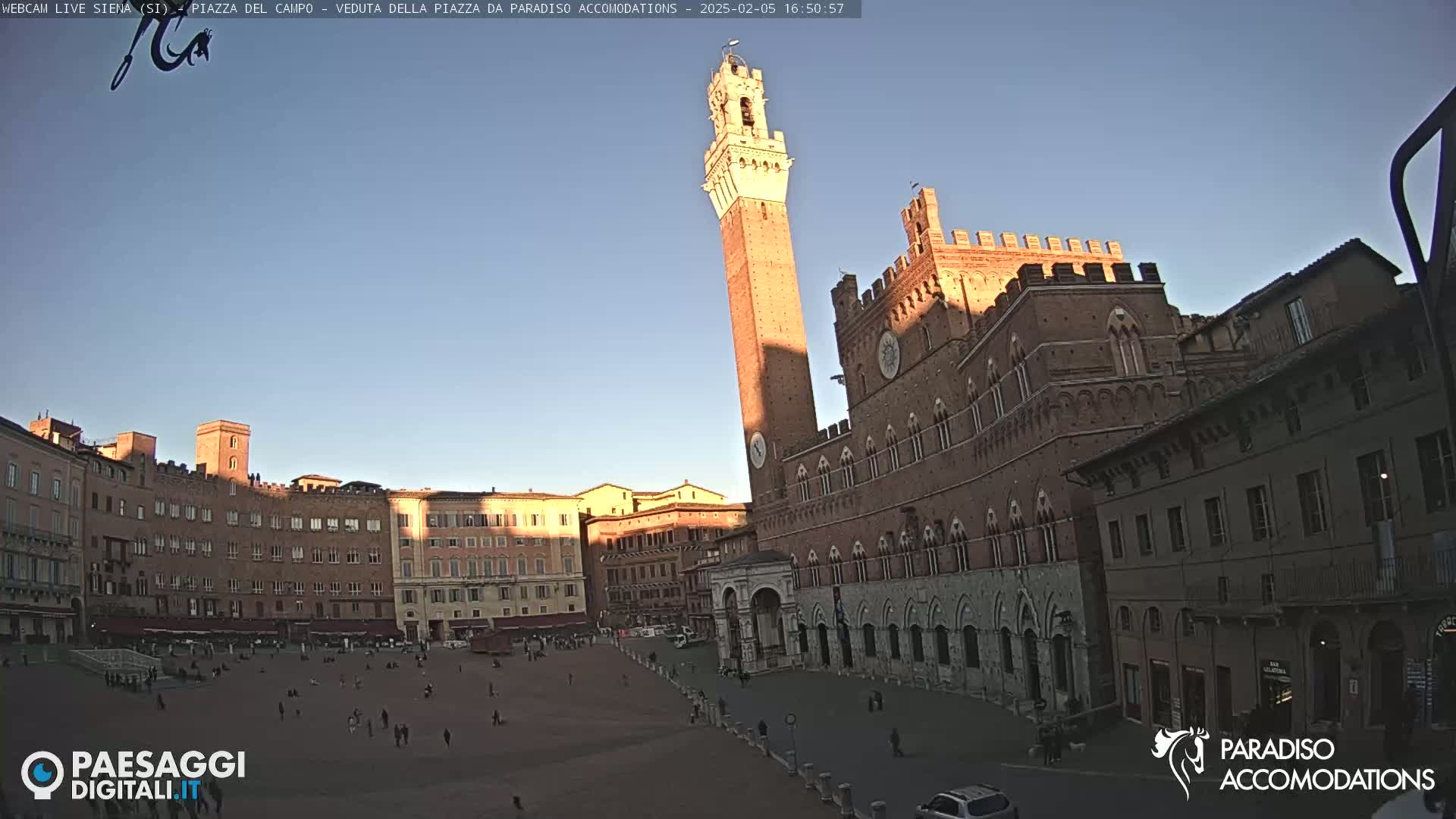 A large, brick piazza in the late afternoon sun, with a tall tower and palatial buildings surrounding a sparsely populated public square.