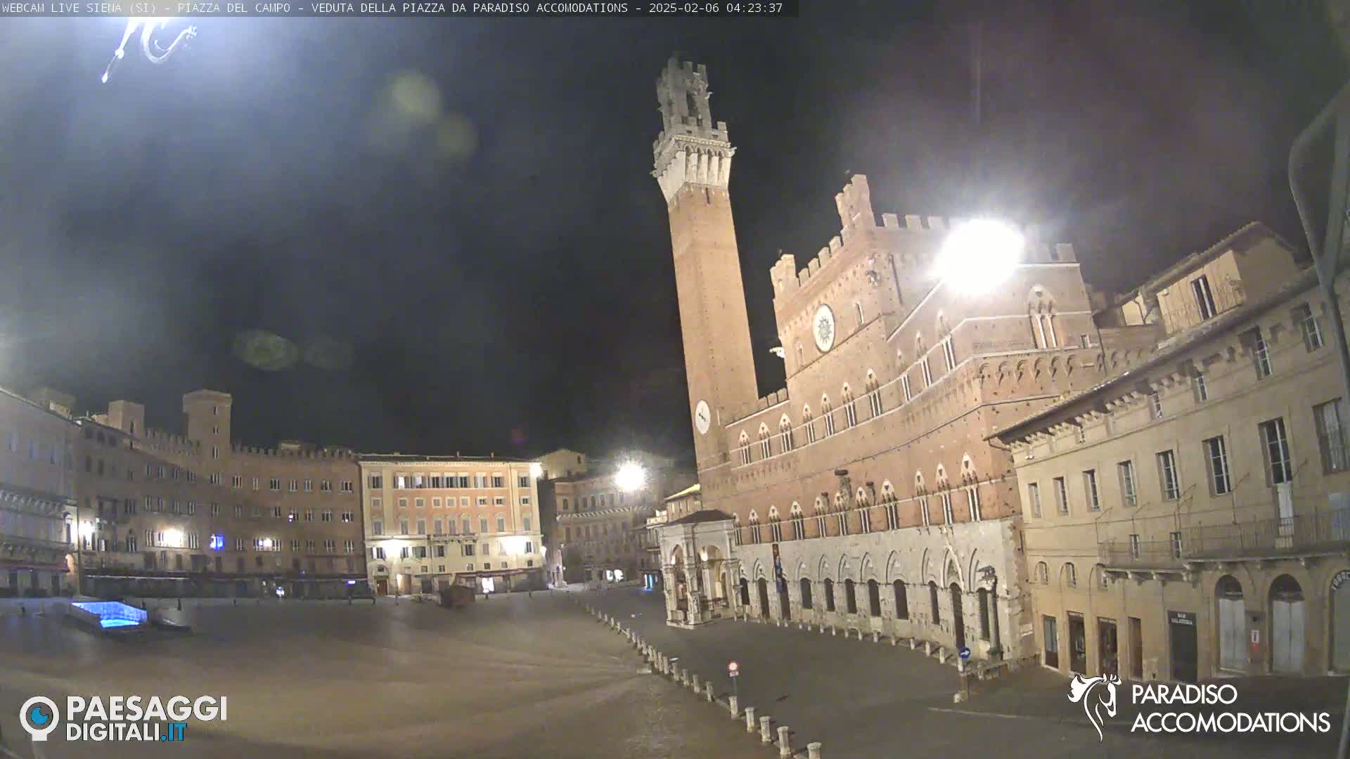 A nighttime view of a large, empty plaza in a city with several historic brick buildings, one prominently featuring a tall tower.