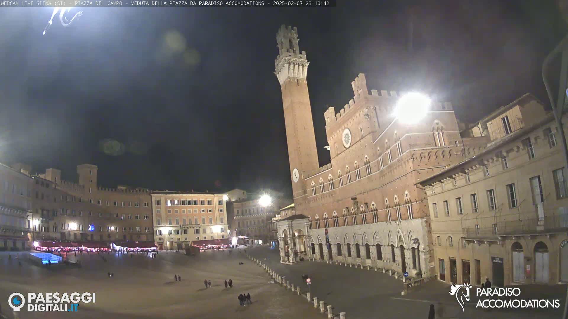 A large, empty town square at night is surrounded by historic brick buildings, including a tall tower, under a dark, clear sky.