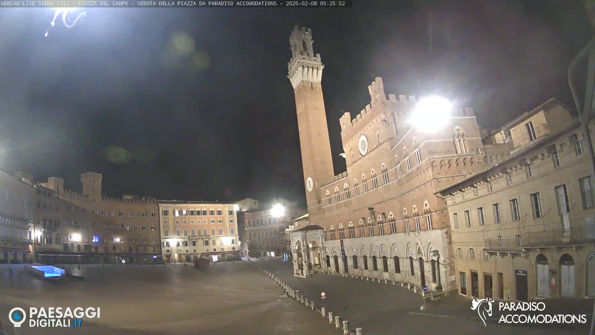 A nighttime view of a large, mostly empty, town square in Italy, featuring a tall brick tower and surrounding buildings under a dark sky.