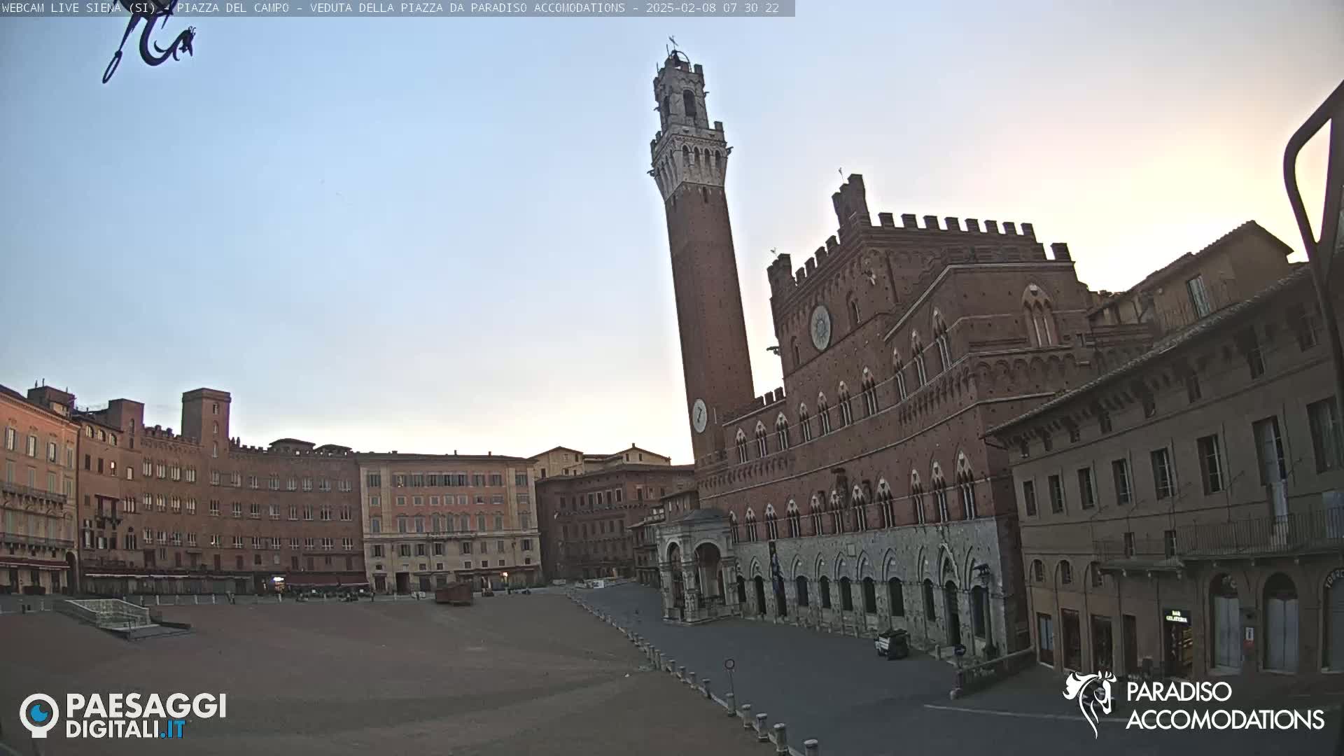 A large, empty plaza is surrounded by medieval-style brick buildings, including a tall tower, under a clear, pale-blue sky.