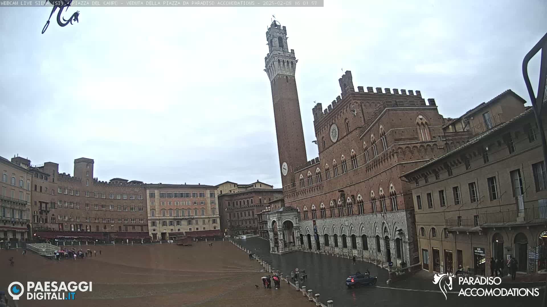A large, brick plaza in a European city on a cloudy day, featuring a tall, slender tower and a grand, arched building.