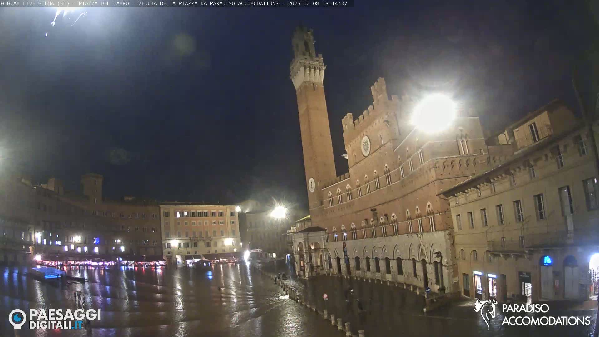 A nighttime view of a flooded town square in the rain, featuring a tall, brick tower and several historic buildings illuminated under dark skies.