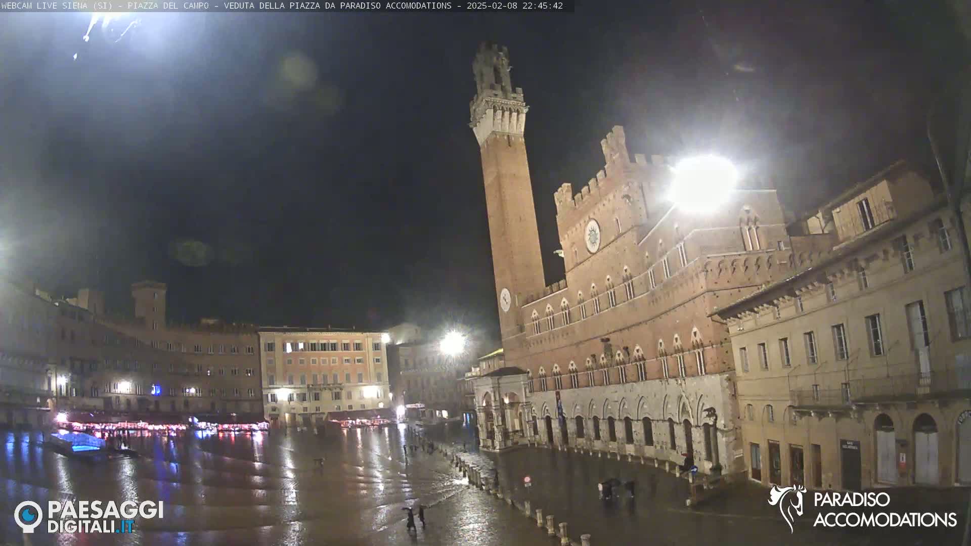 A nighttime, rain-soaked piazza in a European city features a large, brick building with a tall tower, surrounded by other historic structures.