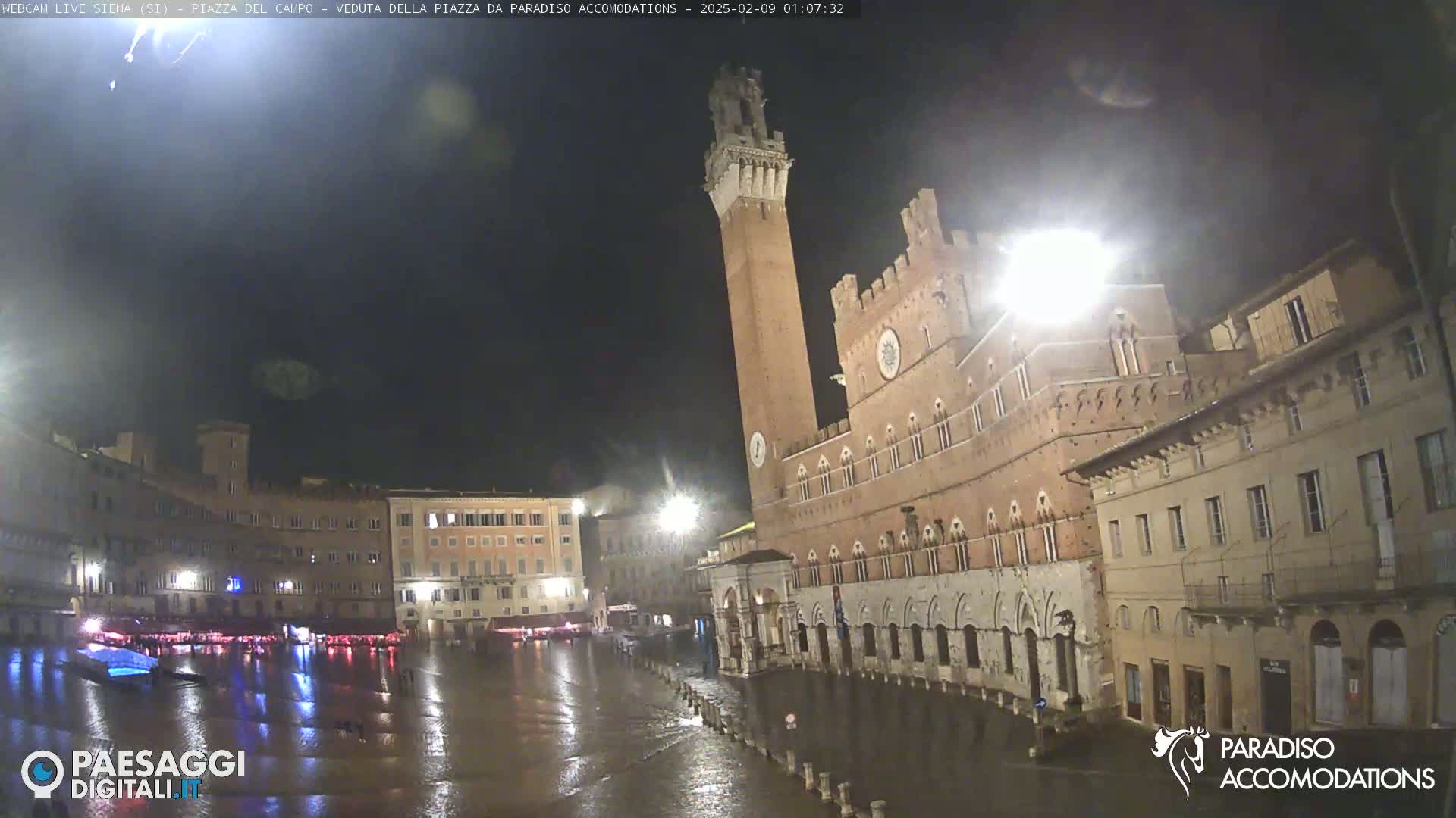 A rain-soaked plaza at night, featuring a tall brick tower and adjacent buildings, is illuminated by streetlights.