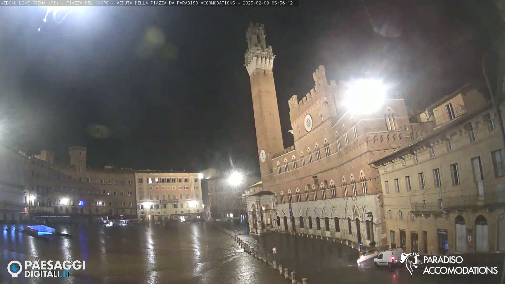 A rain-soaked plaza at night, dominated by a tall brick tower and surrounding medieval buildings under dark skies.