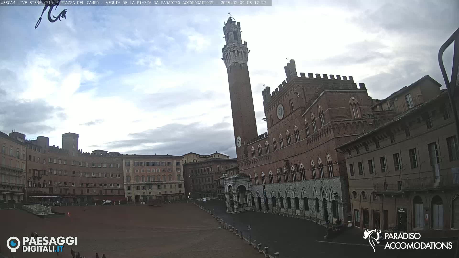 A large, brick piazza in Italy, under a mostly cloudy sky, is dominated by a tall, slender tower and a grand, palatial building.
