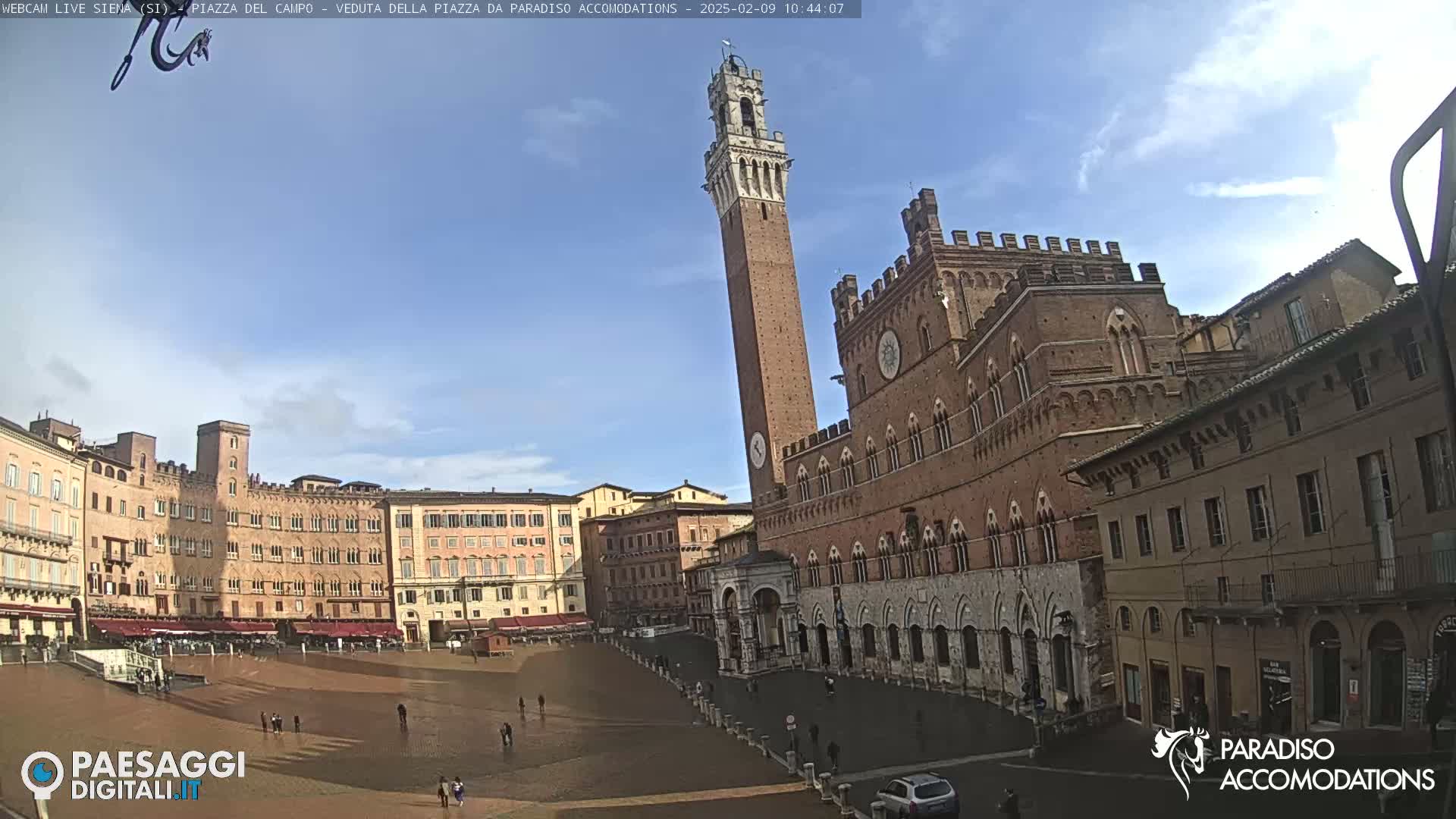 A mostly sunny day reveals a large, brick piazza in Siena, Italy, with a tall tower and several historical buildings surrounding a paved square where a few people are walking.