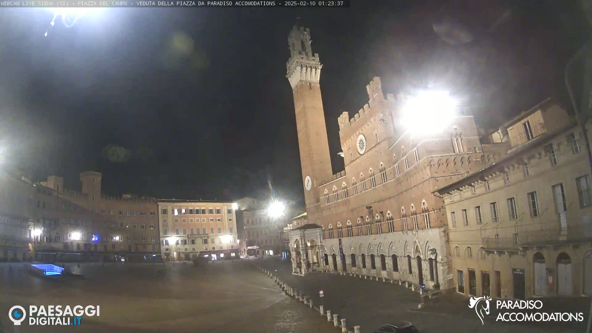 A nighttime view of a large, mostly empty town square in front of a tall, brick tower and adjacent buildings under a dark, slightly hazy sky.