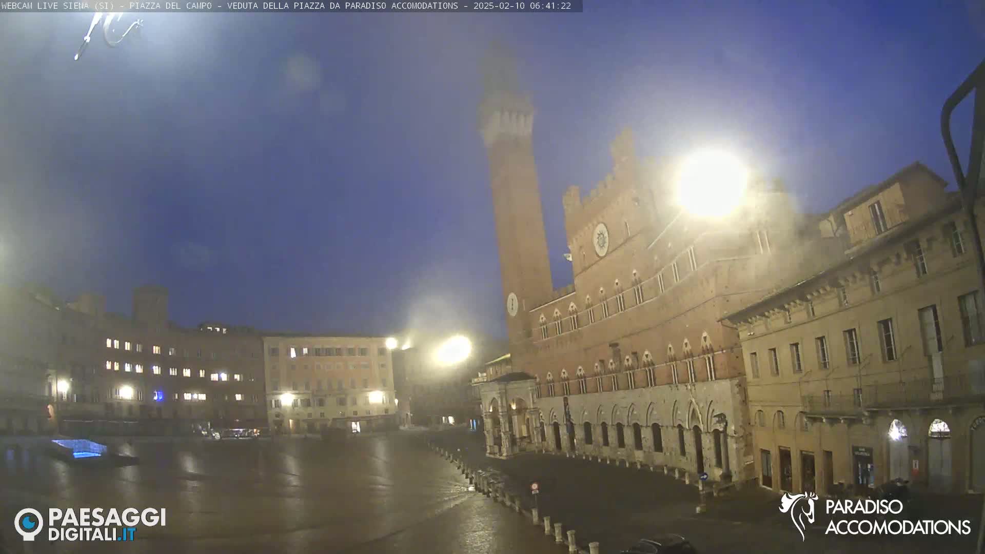 A wet, dimly lit town square at night is surrounded by historic buildings, one of which has a tall tower.