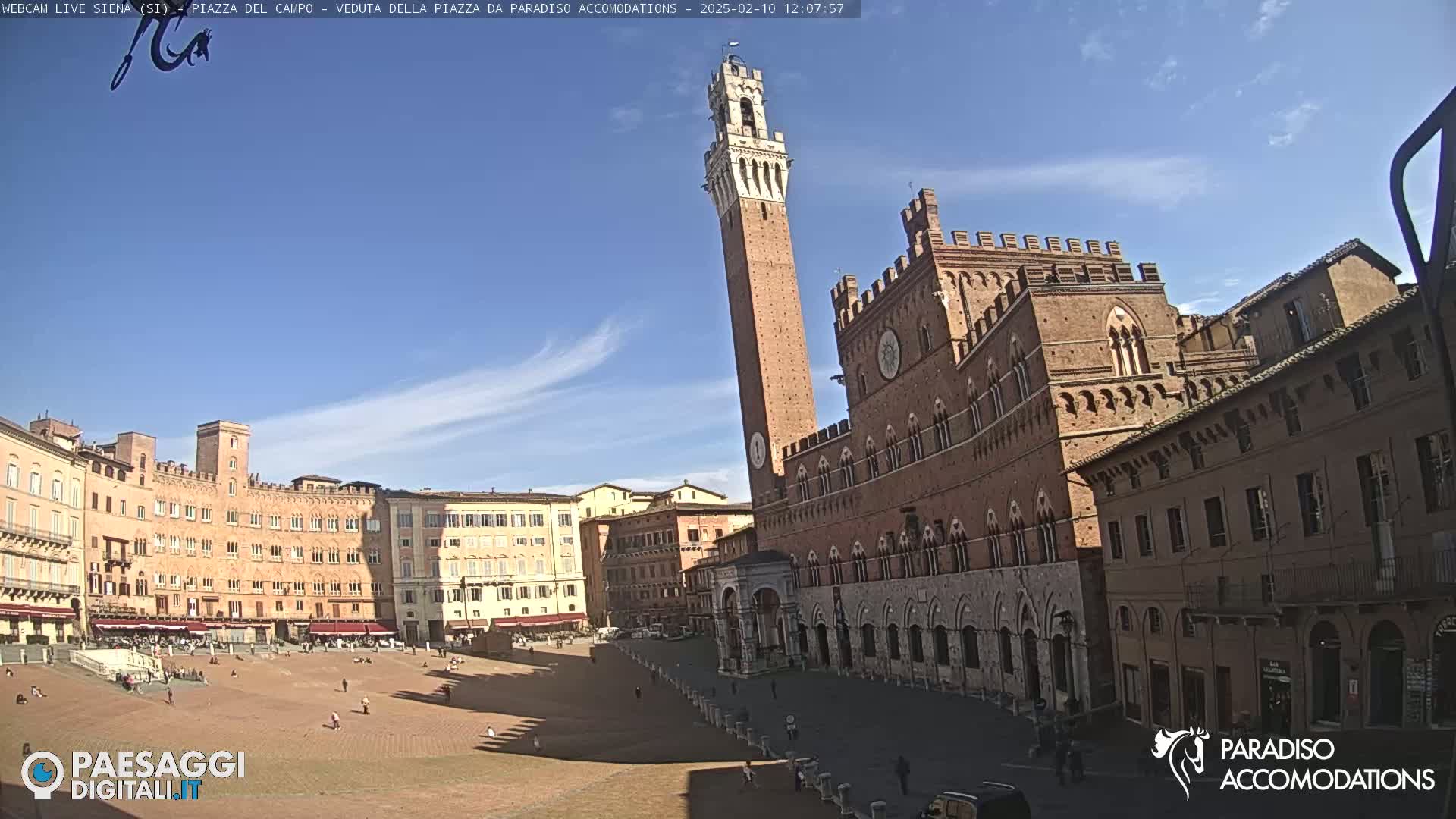 A large, brick piazza in Siena, Italy, is shown on a sunny day with a tall tower and several historic buildings surrounding a large open space.