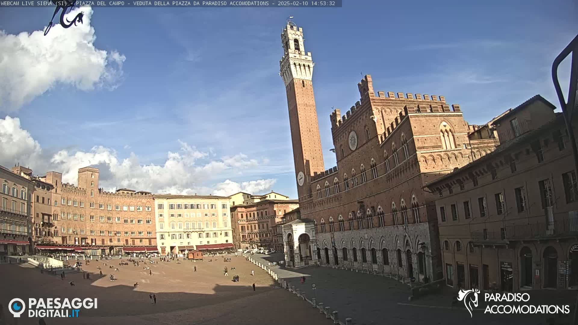 A mostly sunny day shows a large, brick piazza in Italy, with a tall tower and a large, ornate building dominating the scene.