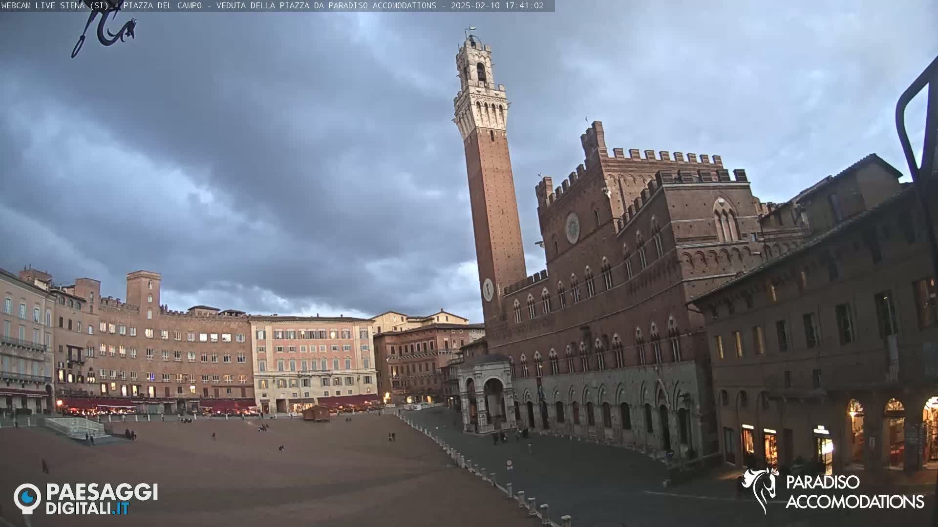 A large, brick piazza in Italy is mostly empty under a cloudy, twilight sky, with a tall tower and several historic buildings surrounding the square.