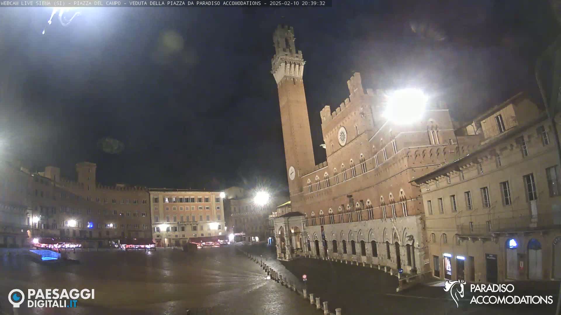 A nighttime view of a large, mostly empty town square in Italy, featuring several historic buildings, including a tall tower, under a dark, cloudy sky.