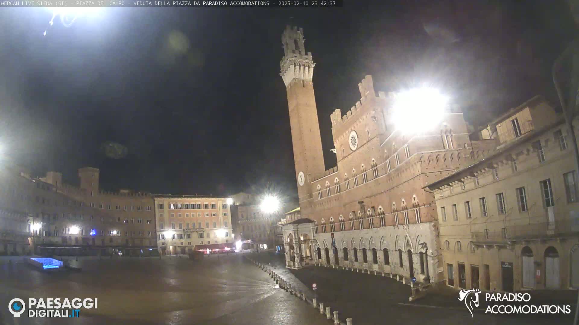 A nighttime view of a large, brick piazza with a tall tower and surrounding buildings under a dark, possibly overcast sky.