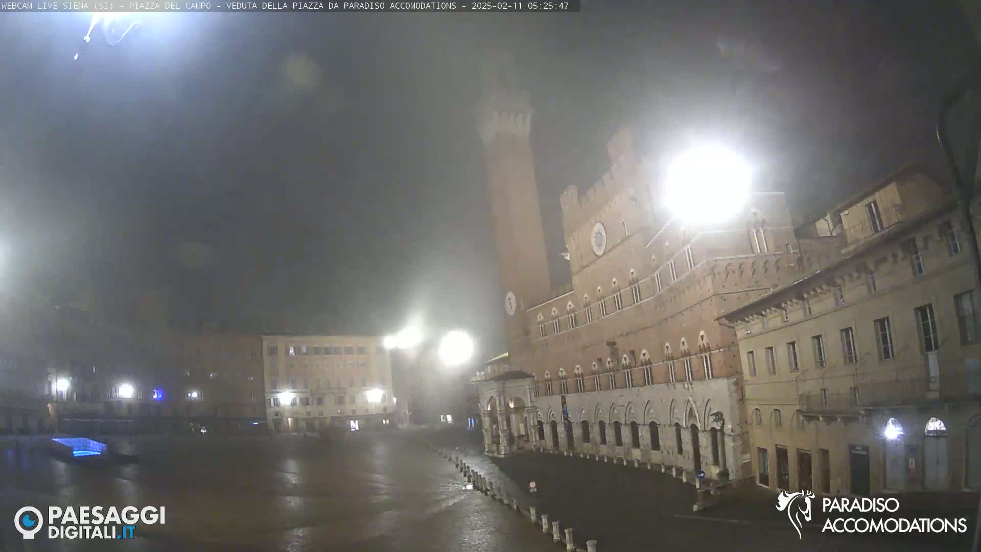 A mostly empty, rain-slicked plaza at night, featuring a tall, medieval-style tower and several buildings under a dark, misty sky.