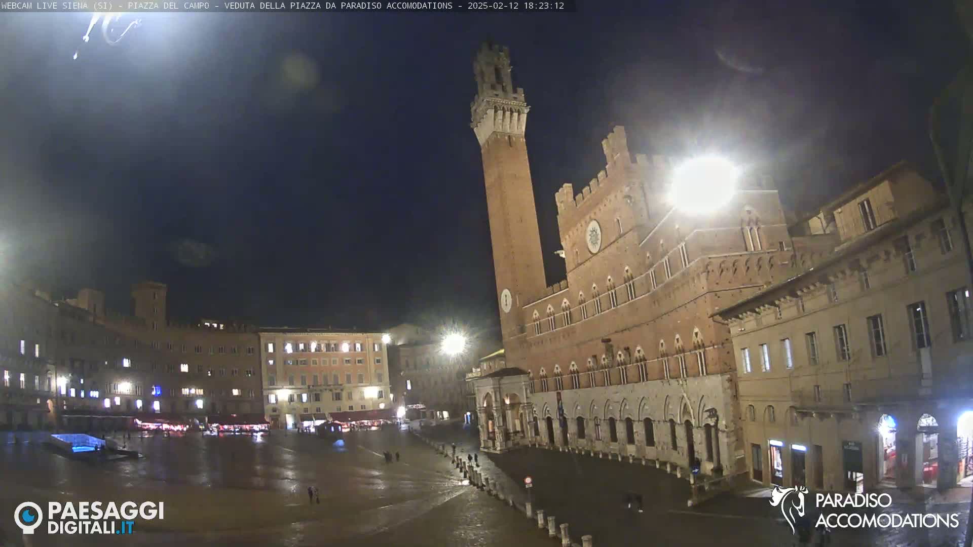 A nighttime view of a large, mostly empty, town square in Italy, featuring a tall brick tower and surrounding medieval-style buildings under a dark sky.