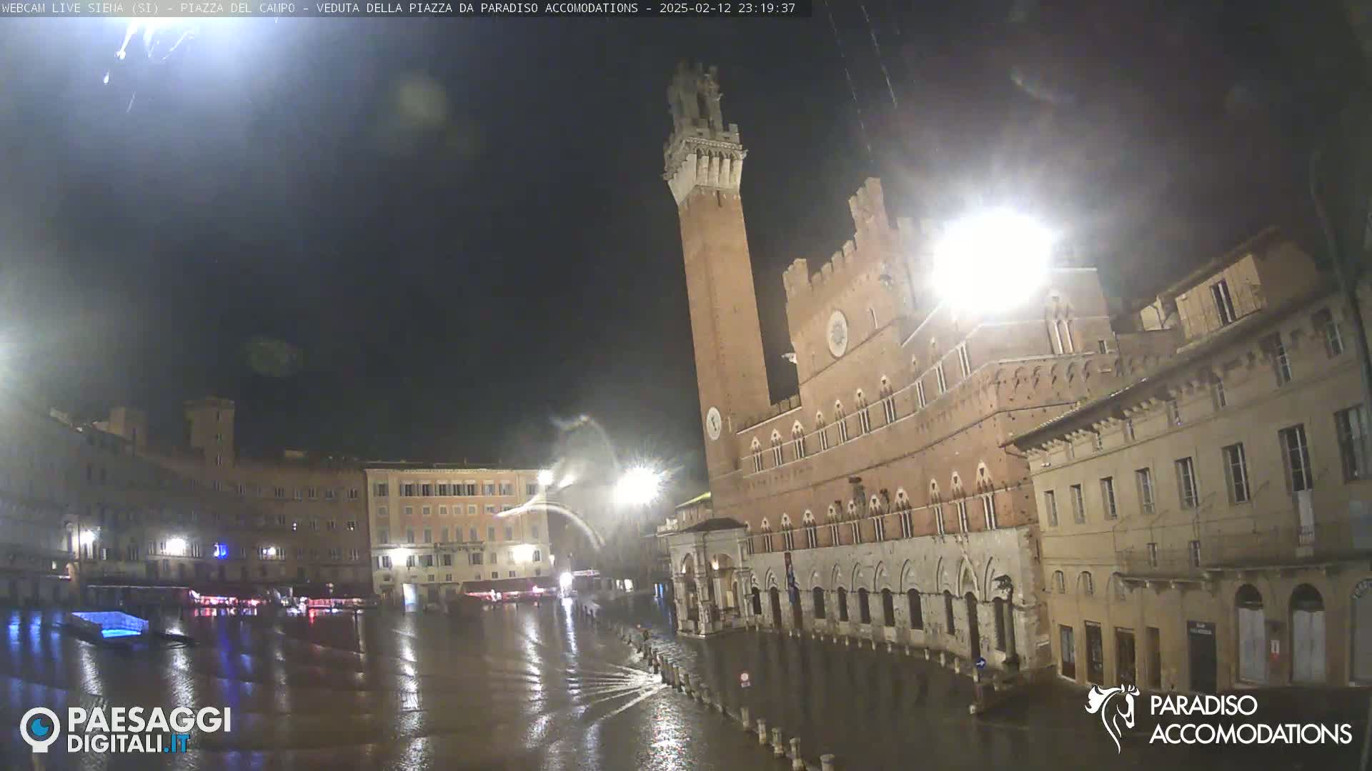 A rain-soaked plaza at night, featuring a tall brick tower and surrounding historic buildings under a dark sky.
