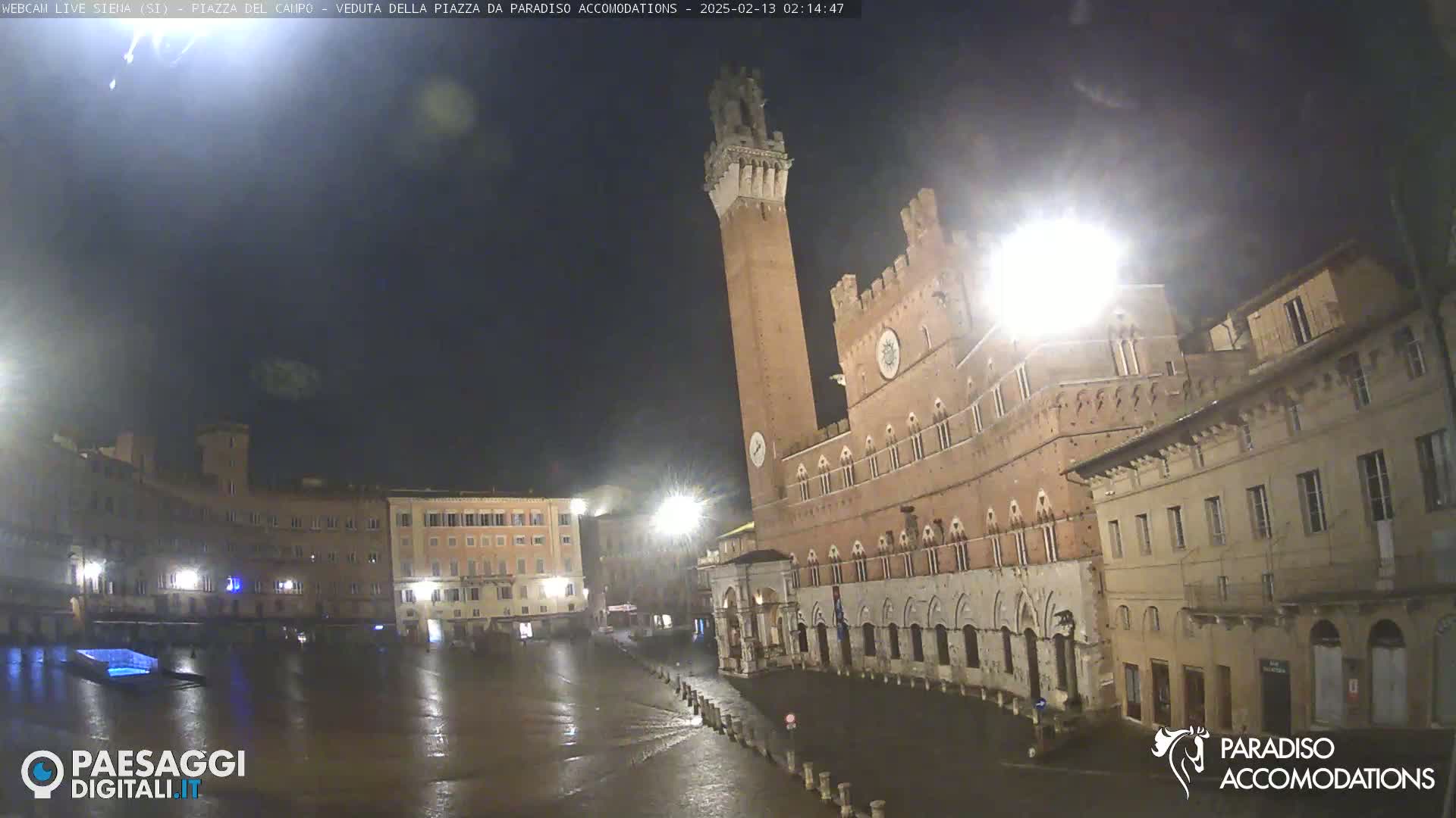 A wet, nighttime view of a large, brick plaza in Italy, featuring a tall tower and several buildings under a dark sky.