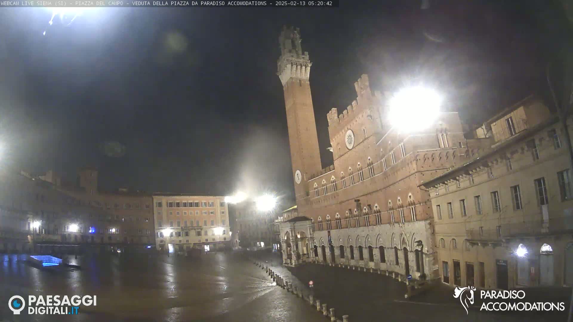 A nighttime view of a large, mostly empty town square in the rain, featuring a tall brick tower and surrounding buildings.