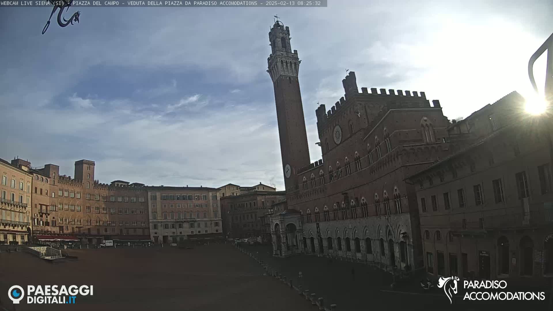 A partly cloudy sky illuminates a large brick piazza in Italy, dominated by a tall tower and medieval buildings.