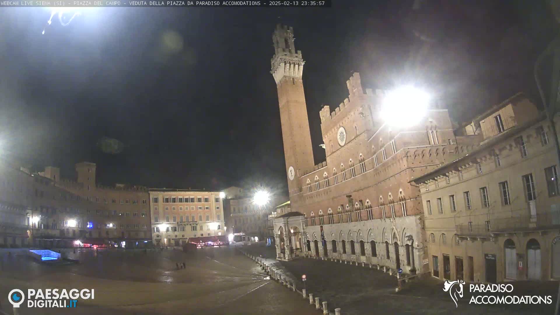 A large, brick piazza at night, illuminated by streetlights, showing a tall tower and surrounding buildings in a wet, empty square.