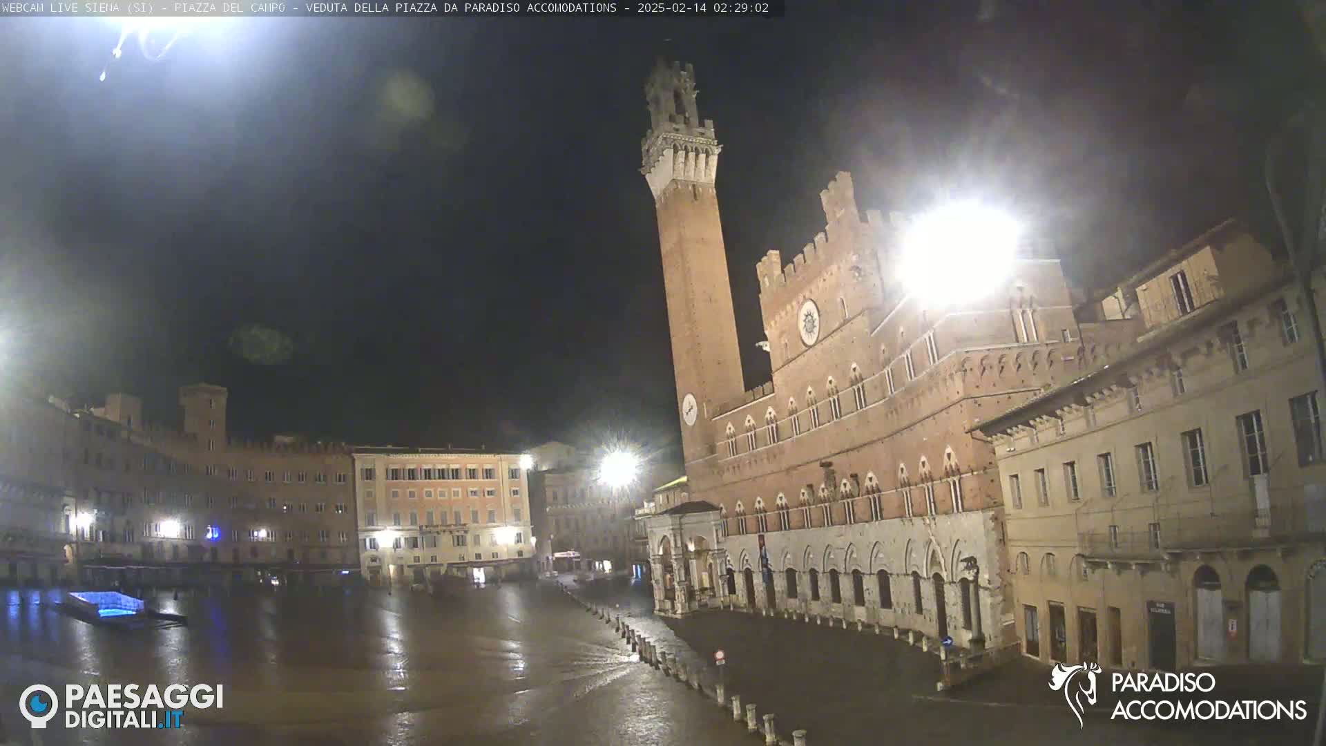 A rain-slicked, nighttime view of a large, empty Italian piazza, dominated by a tall brick tower and adjacent medieval-style buildings.