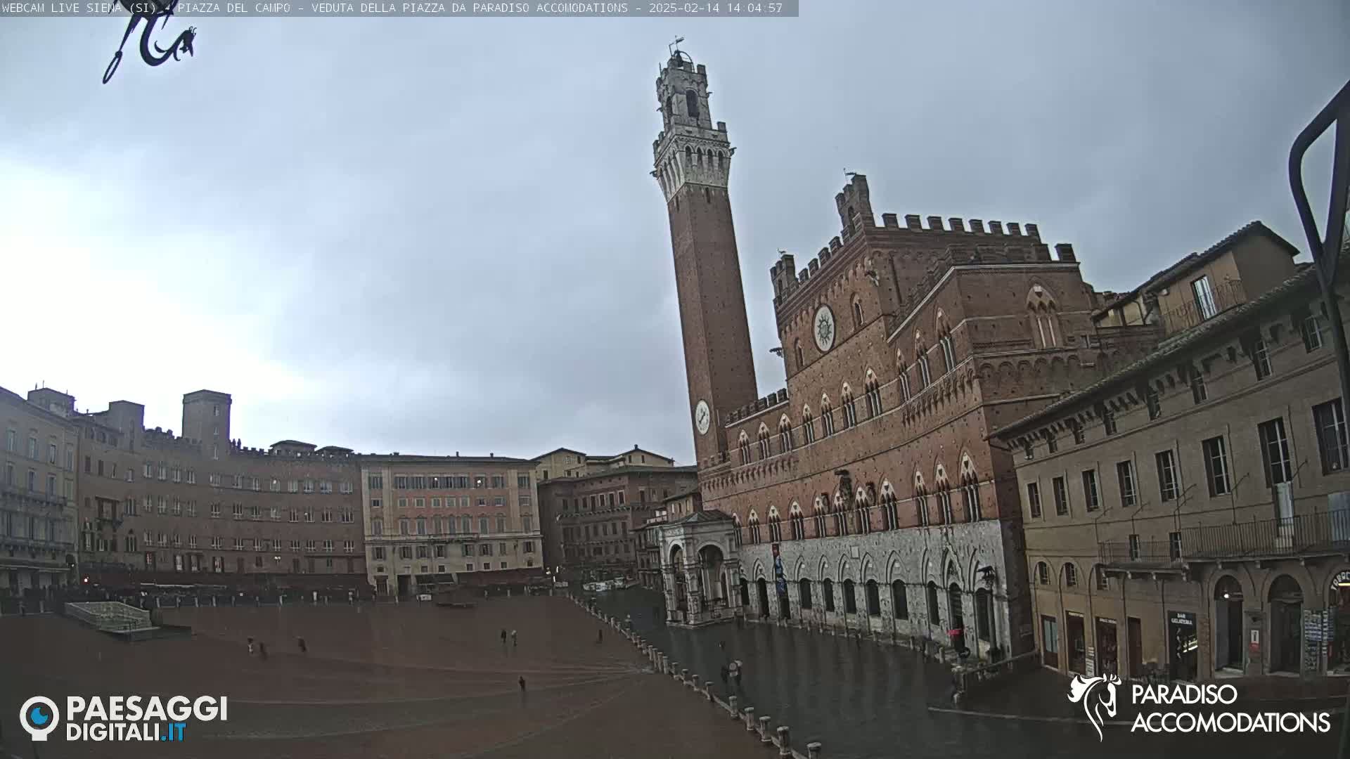 A large, brick public square in a European city is shown on an overcast day with a tall tower and several buildings surrounding it.