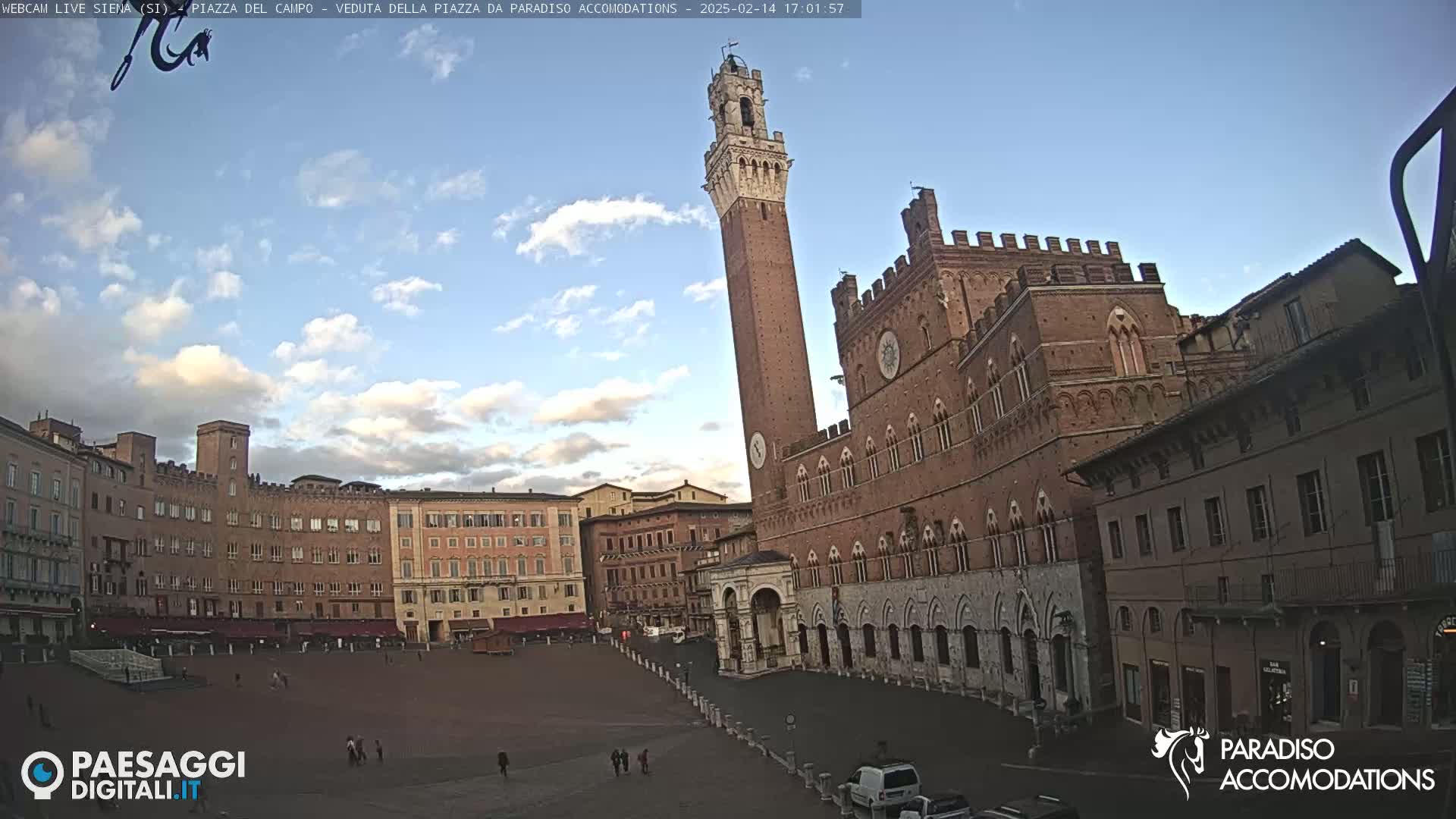 A large, brick public square in Italy, under a partly cloudy sky, is dominated by a tall, slender tower and a grand, brick palazzo.