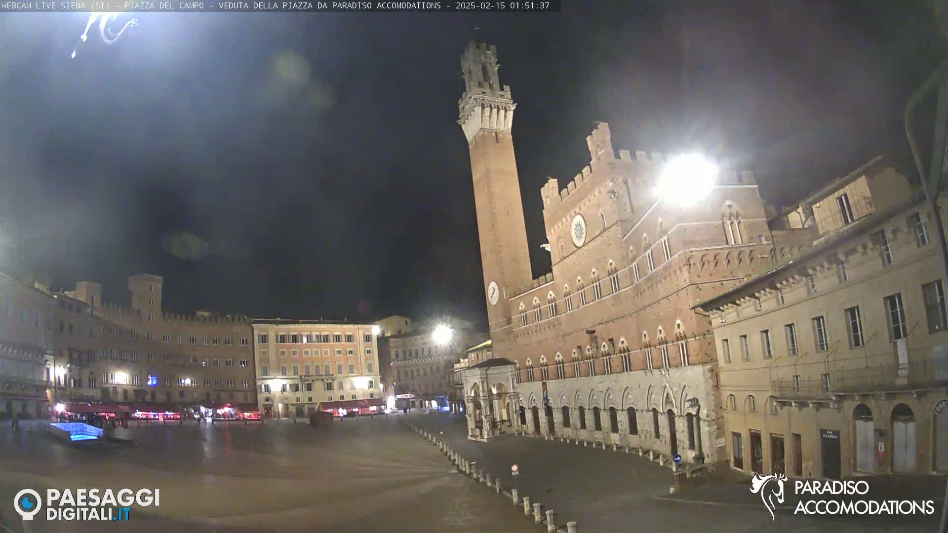 A nighttime view of a large, empty plaza surrounded by historic brick buildings, one prominently featuring a tall tower.