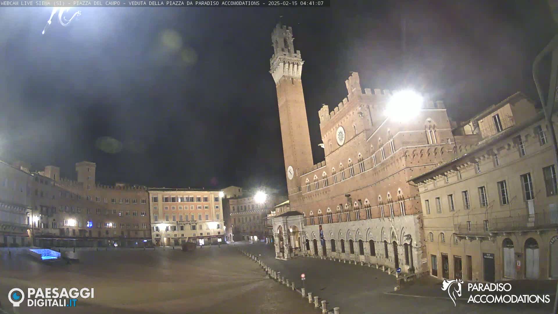 A large, mostly empty, square at night is surrounded by medieval-style buildings, one with a tall tower.