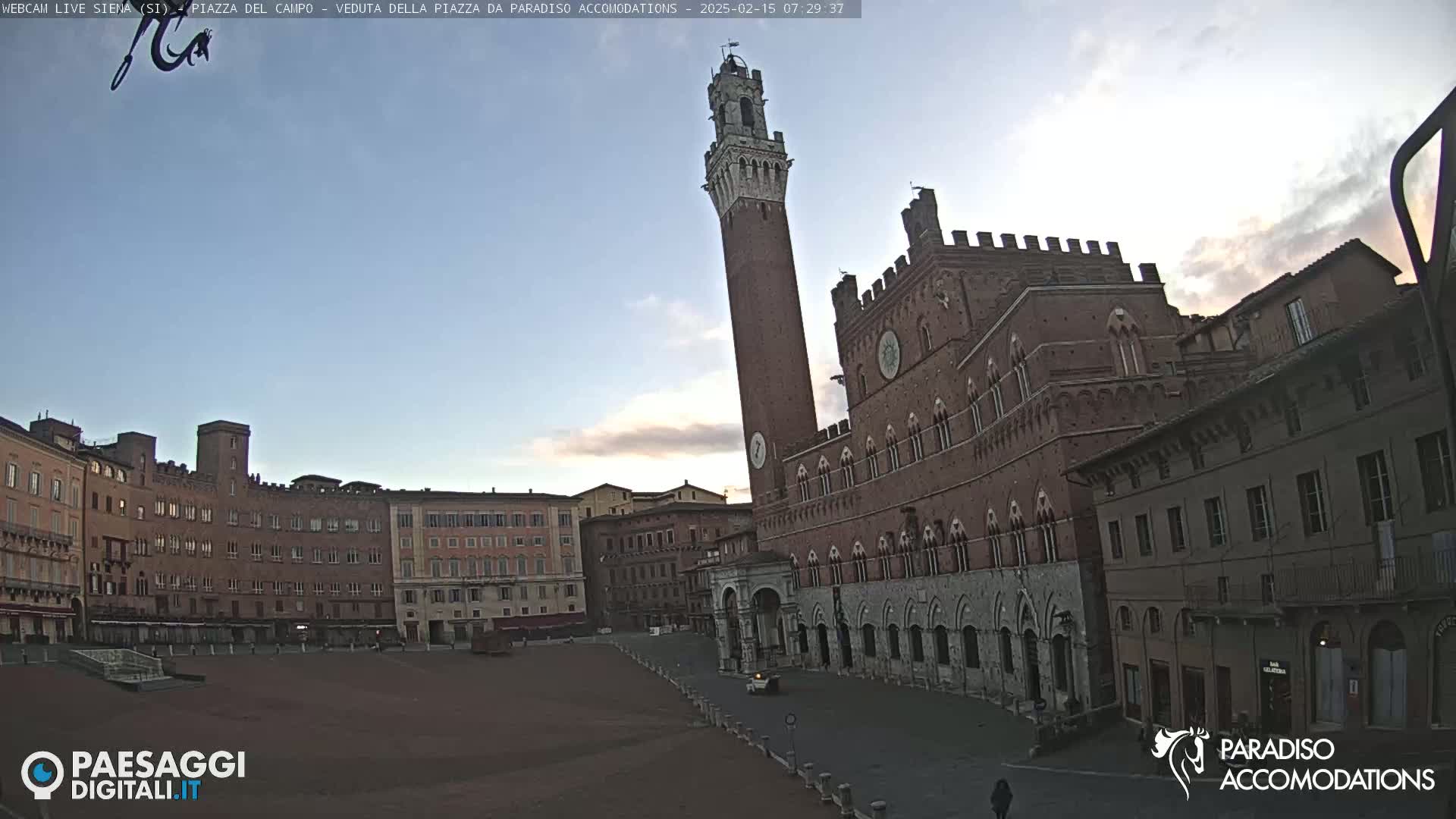 A brick piazza in the early morning light, featuring a tall tower and large palace-like buildings surrounding a mostly empty square.