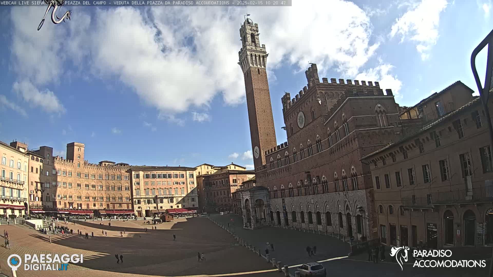 A large, brick piazza in Siena, Italy, is mostly sunny with some clouds, featuring a tall tower and surrounding medieval buildings.