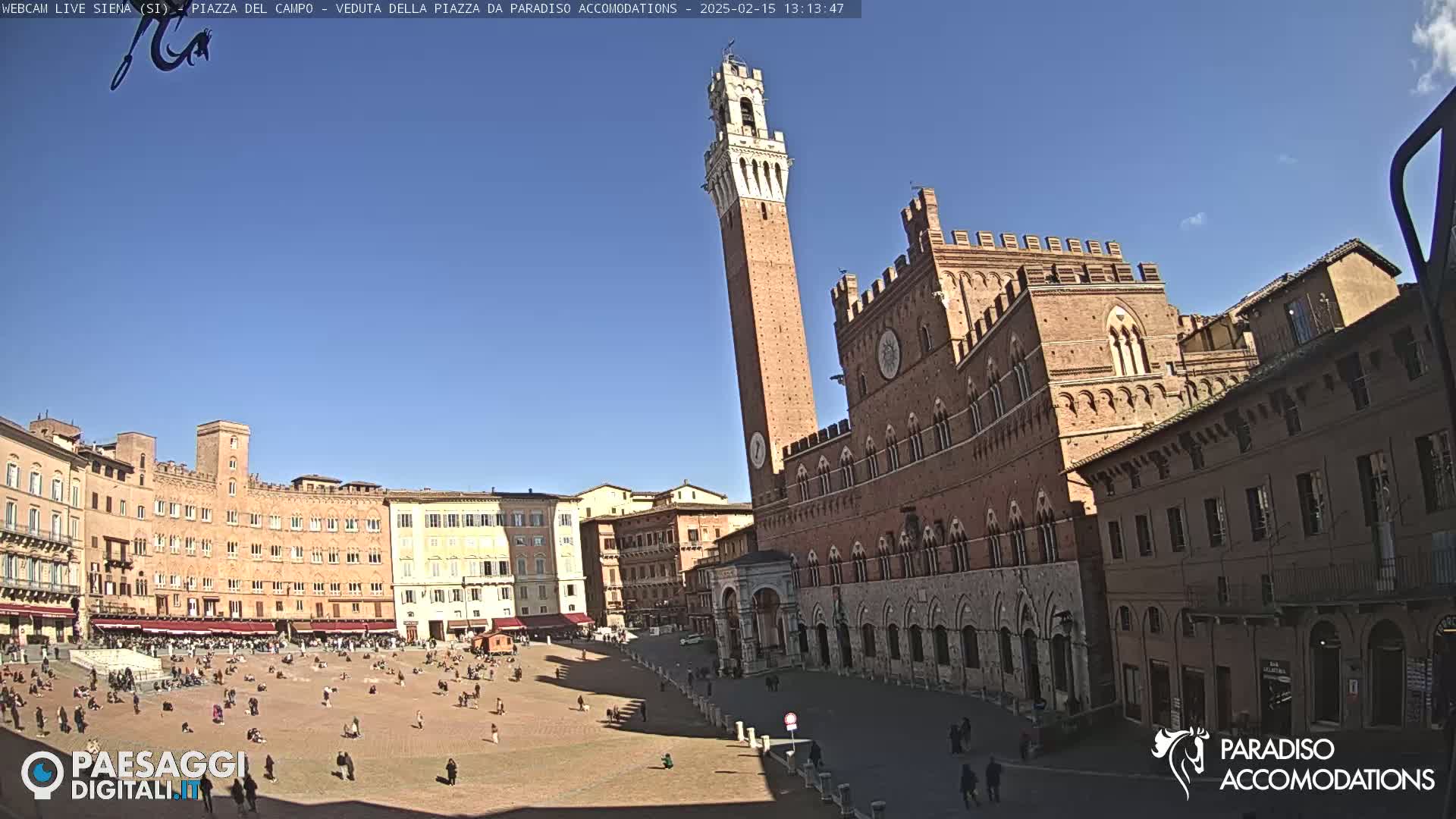 A large, brick piazza in a sunny city is surrounded by historic buildings, including a tall tower and a grand palace.