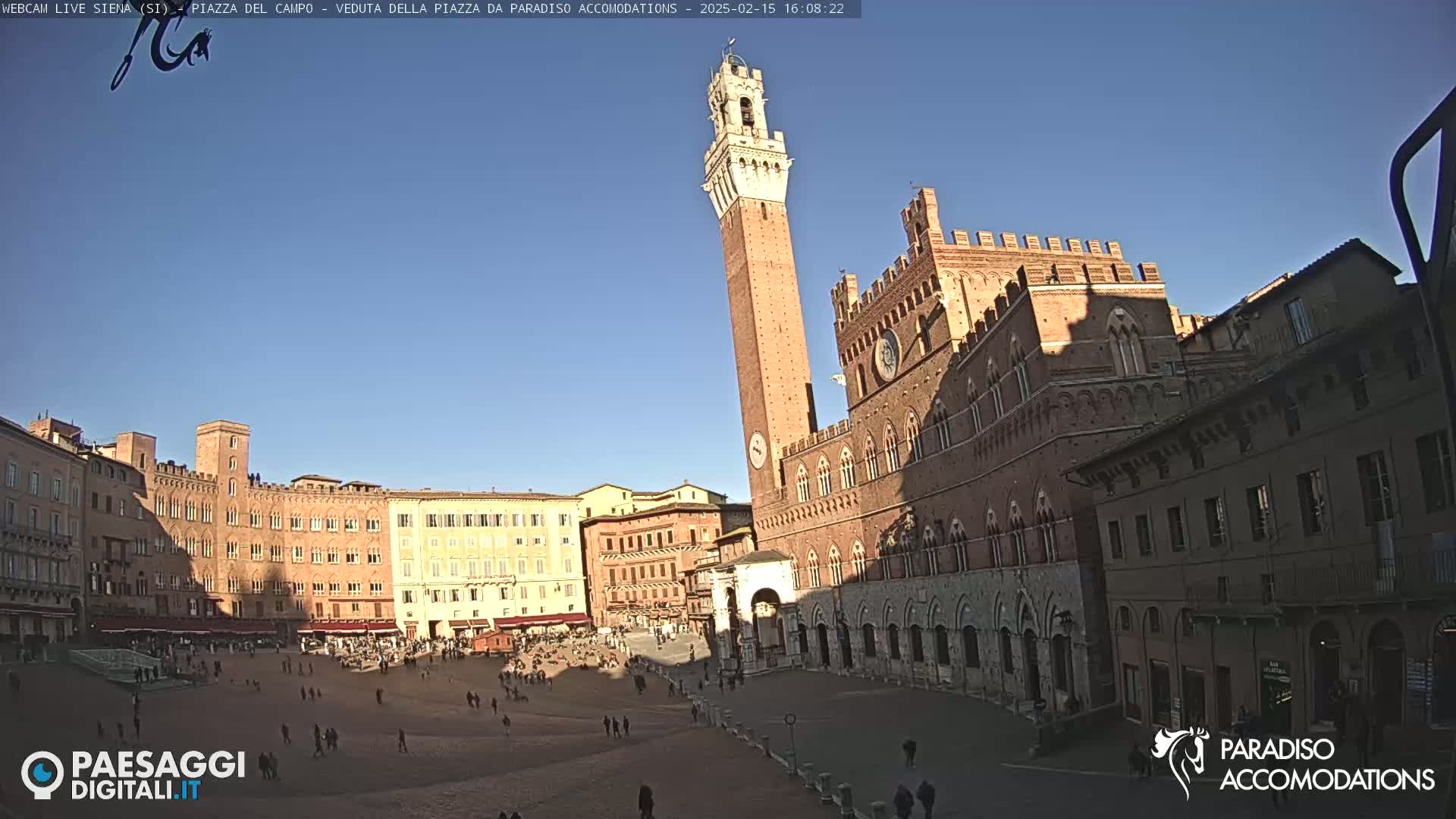 A large, brick piazza in a European city is filled with people under a clear, sunny sky, with a tall, slender tower and imposing brick buildings surrounding the square.