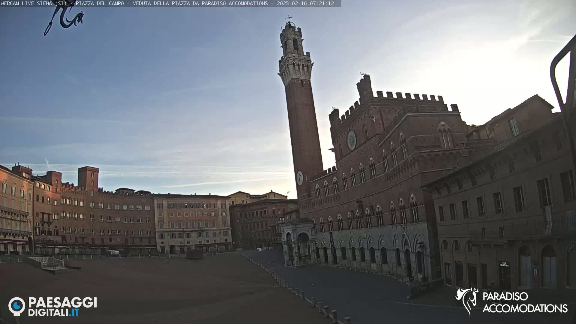 A large, mostly empty, brick plaza is surrounded by medieval-style buildings, including a tall tower, under a clear, light-blue sky.