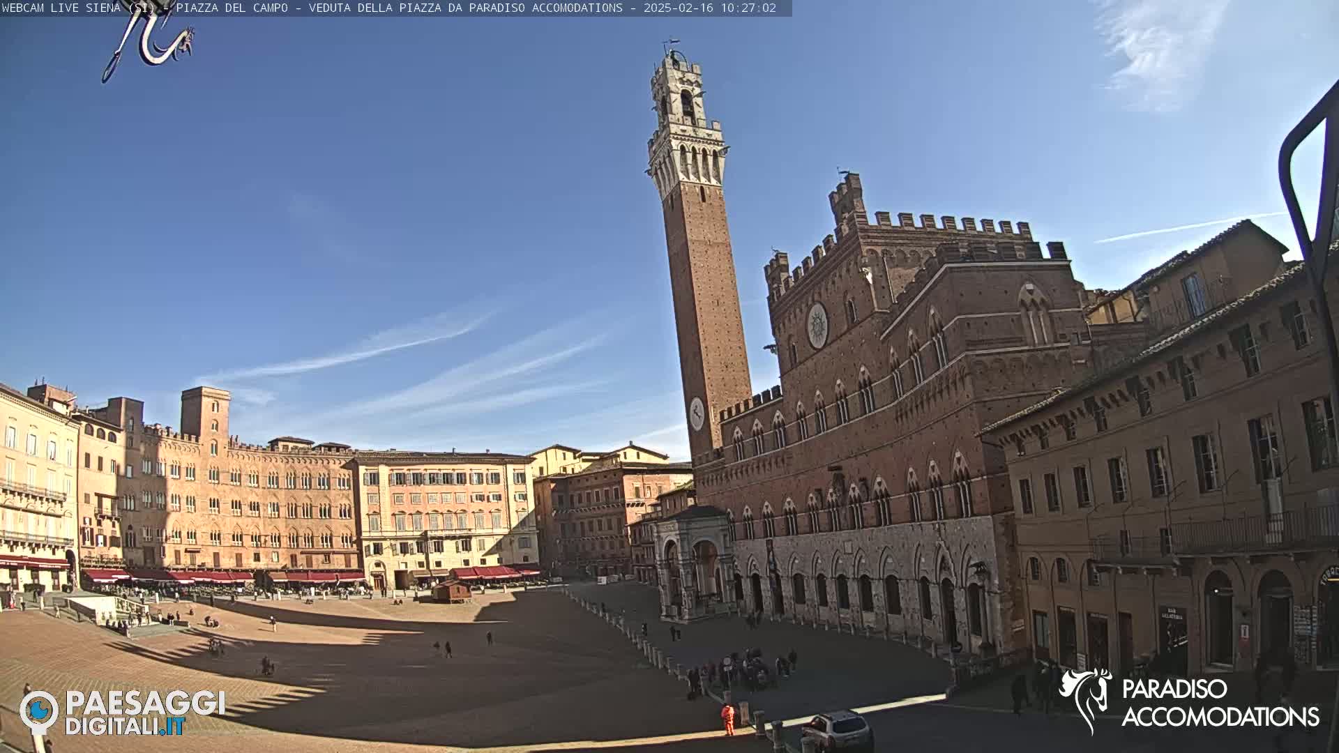 A large, brick piazza in Italy is mostly empty under a clear blue sky, with several historical buildings surrounding it, one prominently featuring a tall tower.