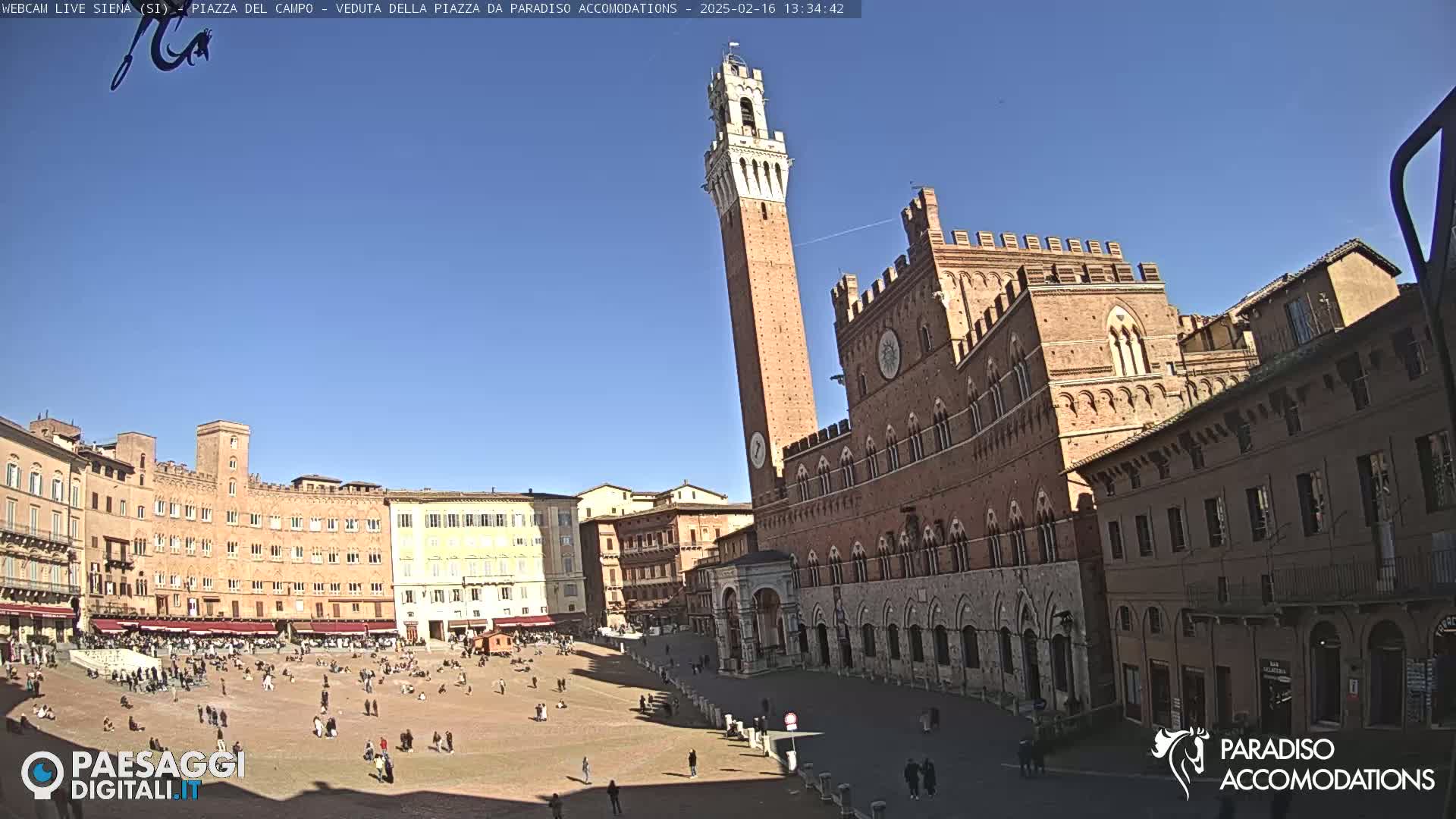 A large, sunny plaza in Italy is surrounded by historic brick buildings, including a tall tower and a palace.