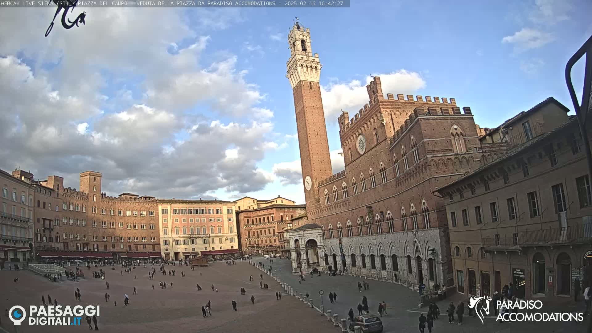 A large, brick piazza in Italy, under a partly cloudy sky, is filled with people and surrounded by historic buildings, including a tall tower.