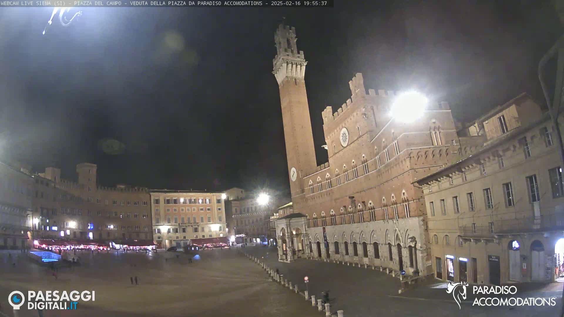 A nighttime view of a large, mostly empty plaza in a European city, with a tall brick tower and surrounding buildings illuminated.