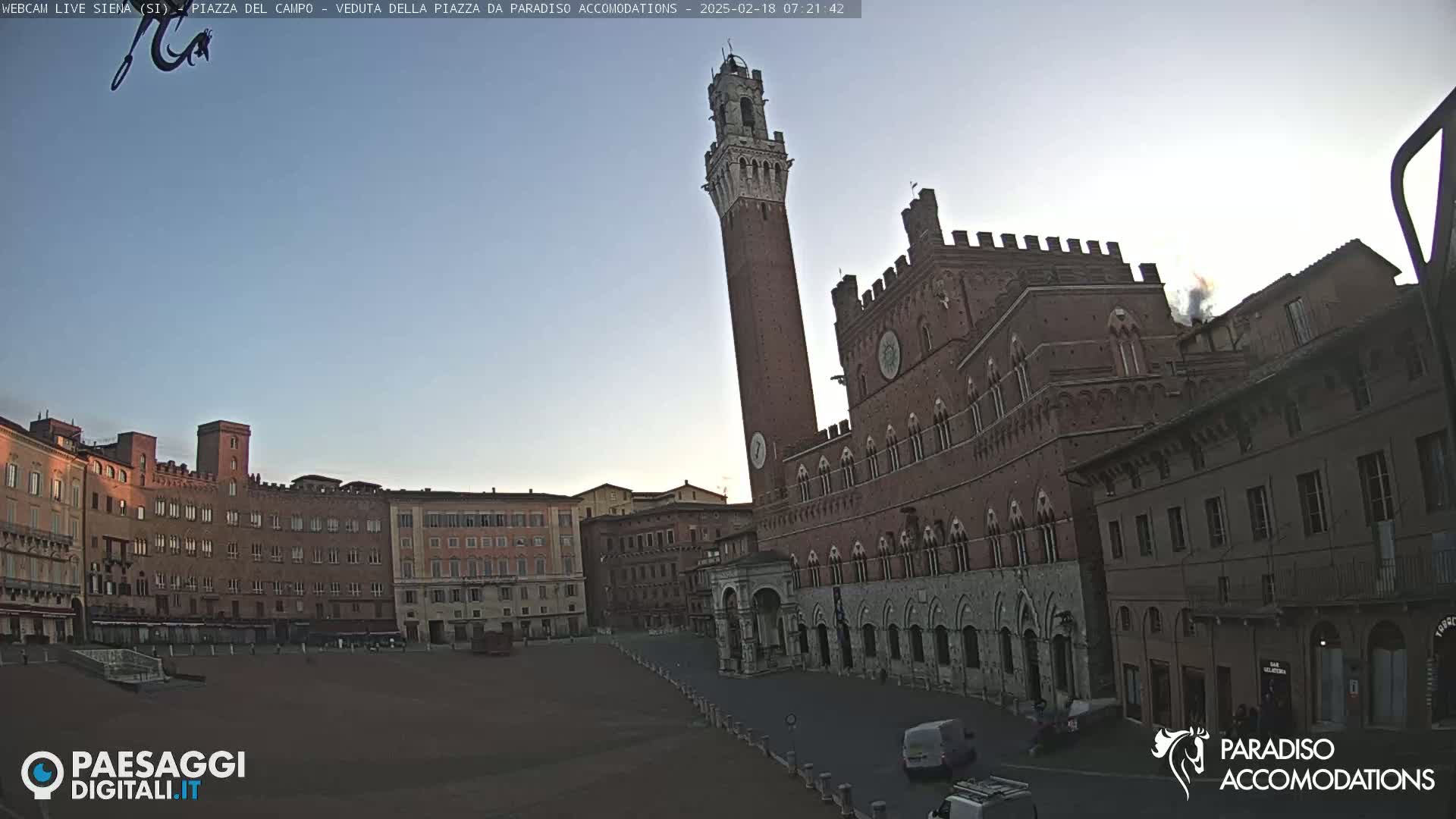 A large, brick piazza in the early morning, with a tall tower and medieval buildings surrounding a mostly empty square under a clear sky.