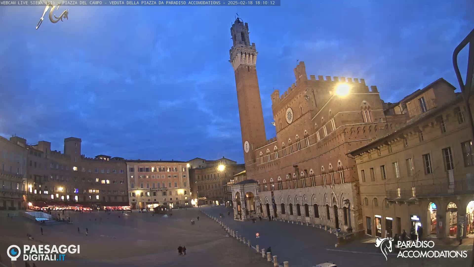 A large, mostly empty, stone plaza at twilight is surrounded by medieval-style buildings, including a tall brick tower.