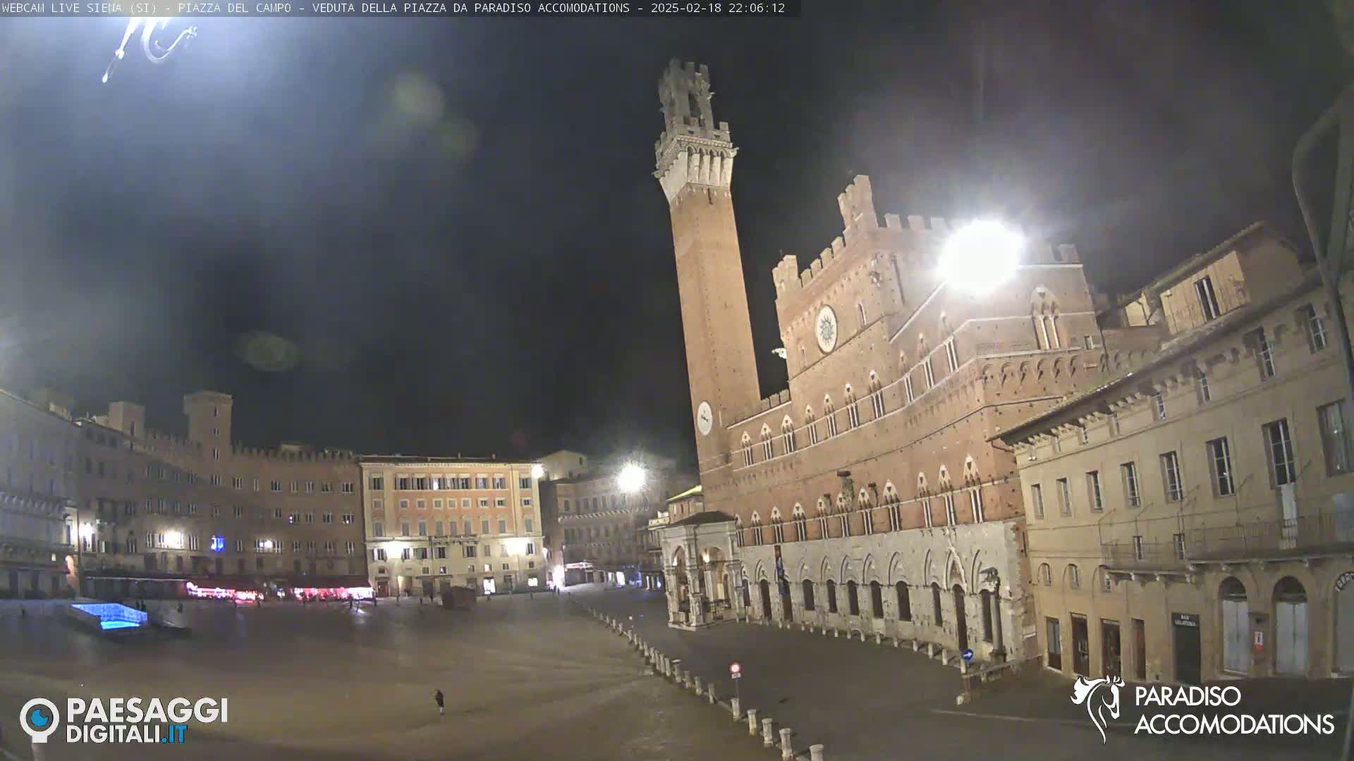 A large, empty plaza at night is surrounded by brick buildings, including a tall tower and a palace-like structure.