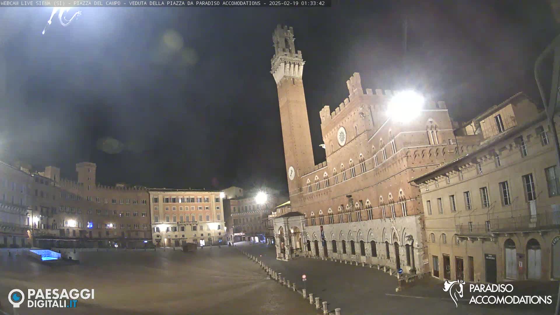 A nighttime view of a large, empty town square in Italy, surrounded by historic brick buildings, one of which has a tall tower.
